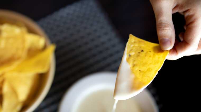 A person dipping a tortilla chip into a bowl of queso, but it’s dripping off the chip because there is too much cheese