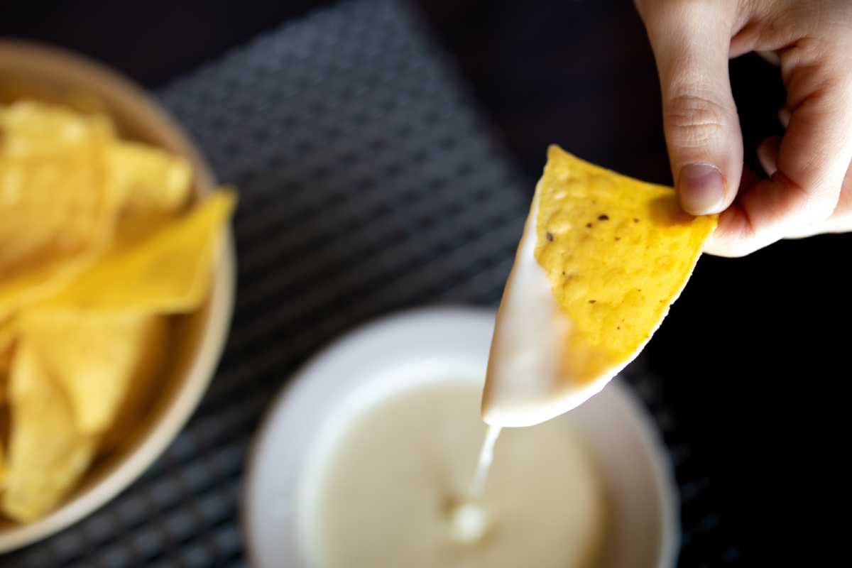 A person dipping a tortilla chip into a bowl of queso, but it’s dripping off the chip because there is too much cheese